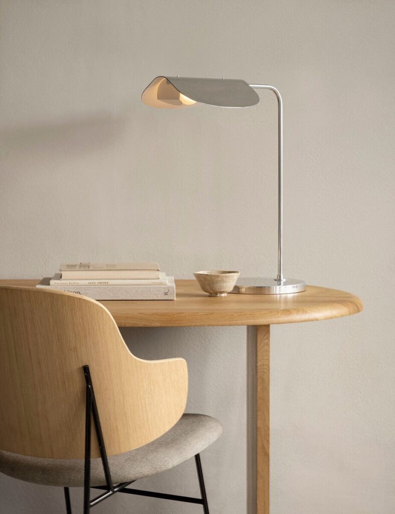 Minimalist Scandinavian-style wooden desk setup with a modern metal desk lamp, a curved wooden chair with fabric seat, a small ceramic bowl, and stacked neutral-toned books against a light beige wall.